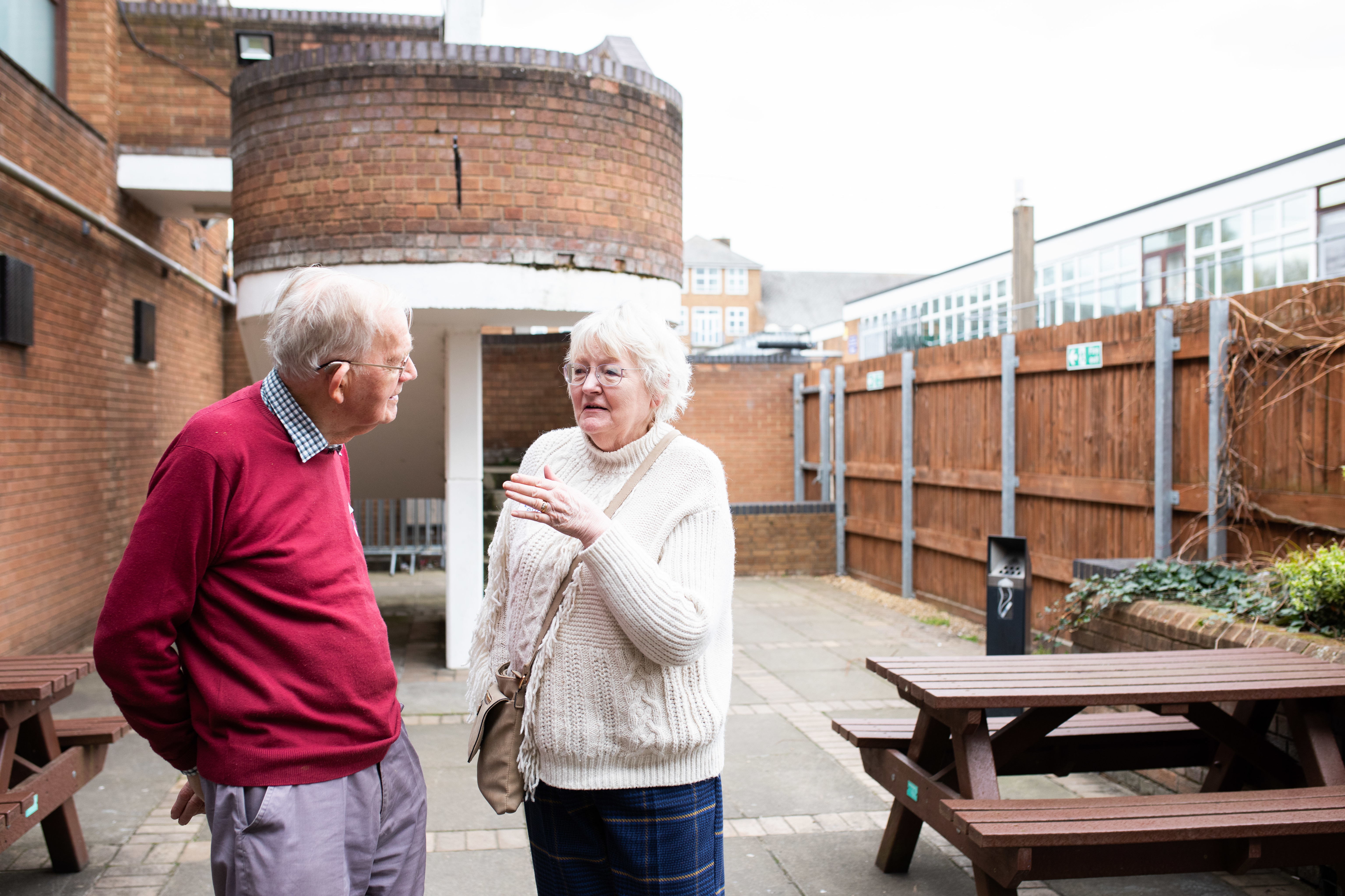 An older man and woman standing in a courtyard with a picnic table in the background. They are talking to each other