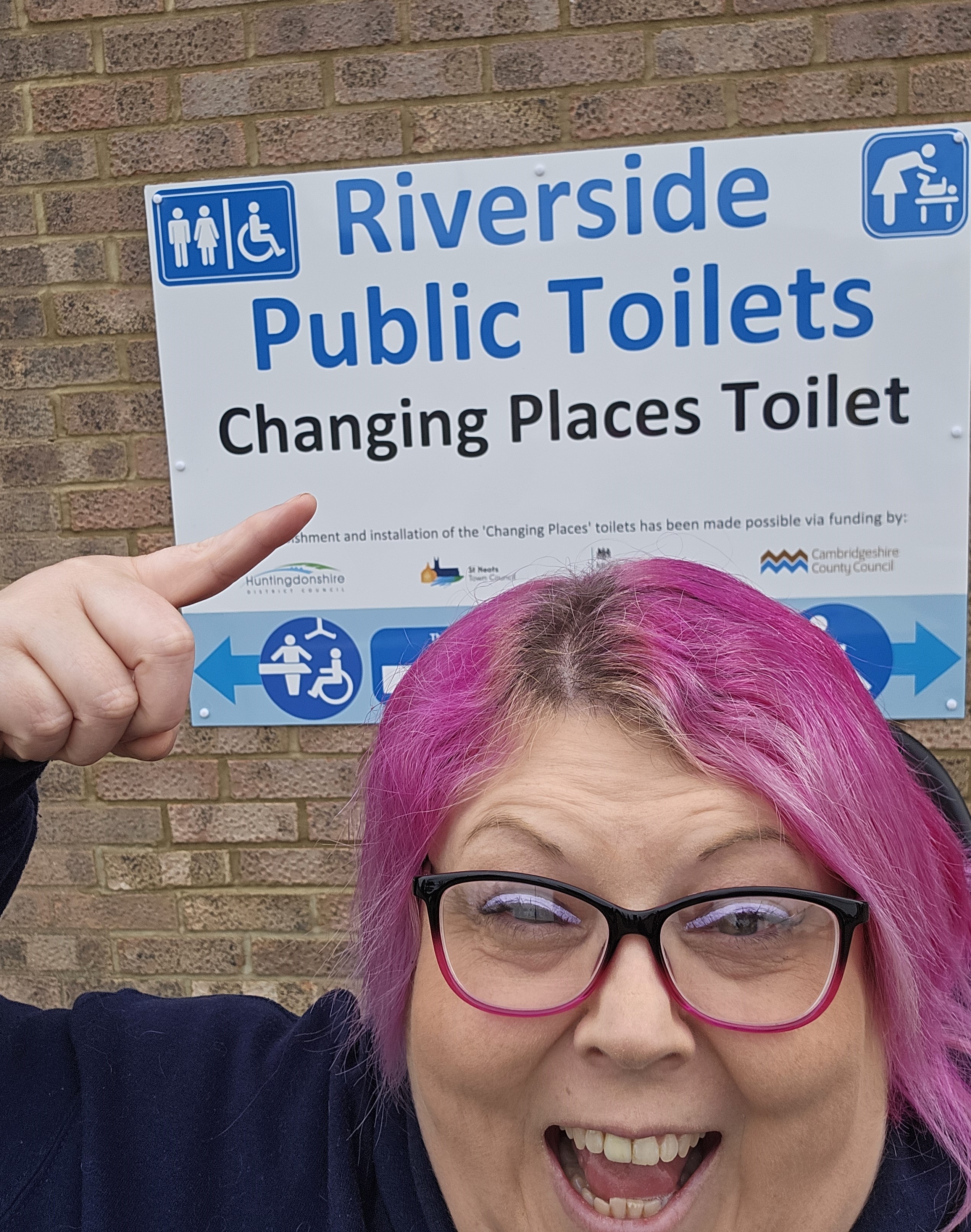 Liz Owen in front of the Changing Places Toilet sign and pointing to it