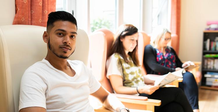 Three people sitting in a room. Two are reading and the other is looking at the camera.