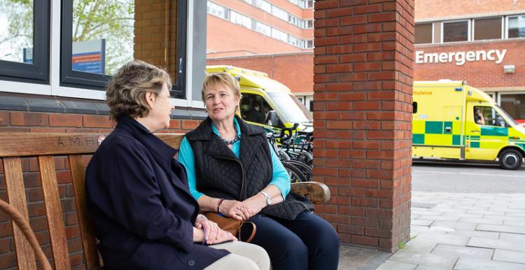 Two women sitting on a bench outside a hospital and talking to each other. There is an ambulance parked in the background.