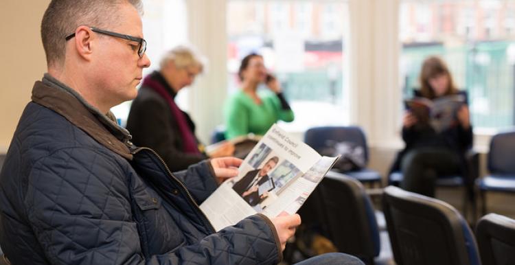 Four people sitting in a surgery waiting room. Three are reading and one is using their mobile phone.