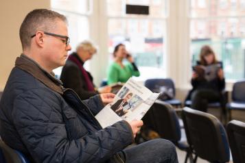 A man sitting in a GP surgery waiting room reading a magazine. Other people are seated in the background reading or using their mobile phones.