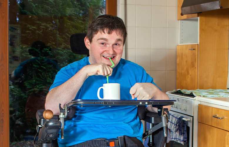 Picture shows man in wheelchair in his kitchen having a drink using a straw